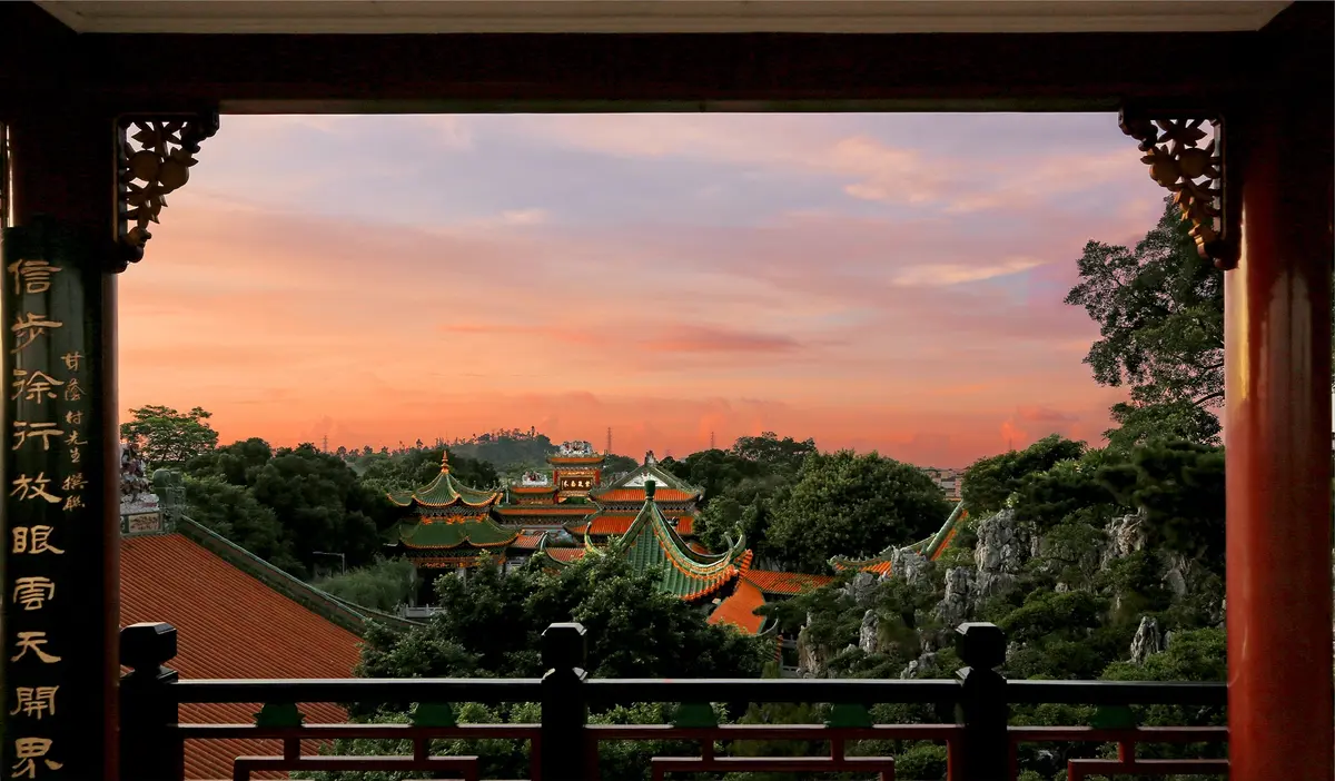 Sunset glow illuminating curved Chinese tiled roofs, stone rockeries and garden pavilions across peaceful water in Panyu