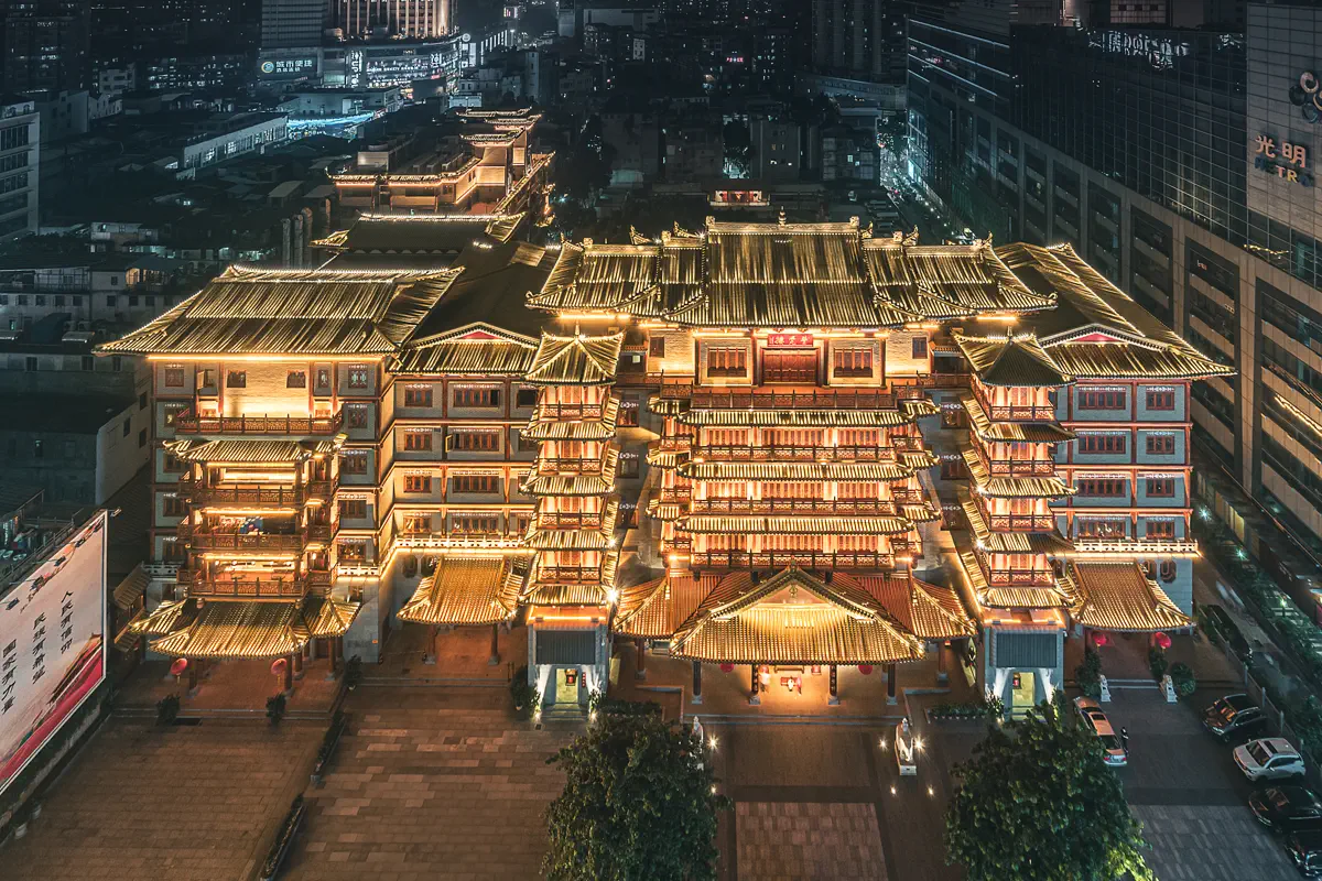 High-angle aerial night view of Dafo Temple's illuminated golden roof pavilions surrounded by modern urban Guangzhou