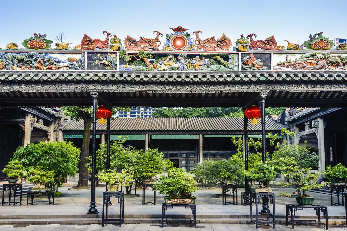 Covered breezeway corridor in Chen Clan Ancestral Hall courtyard with hanging red lanterns, potted Lingnan penjing bonsai, and carved beam frames