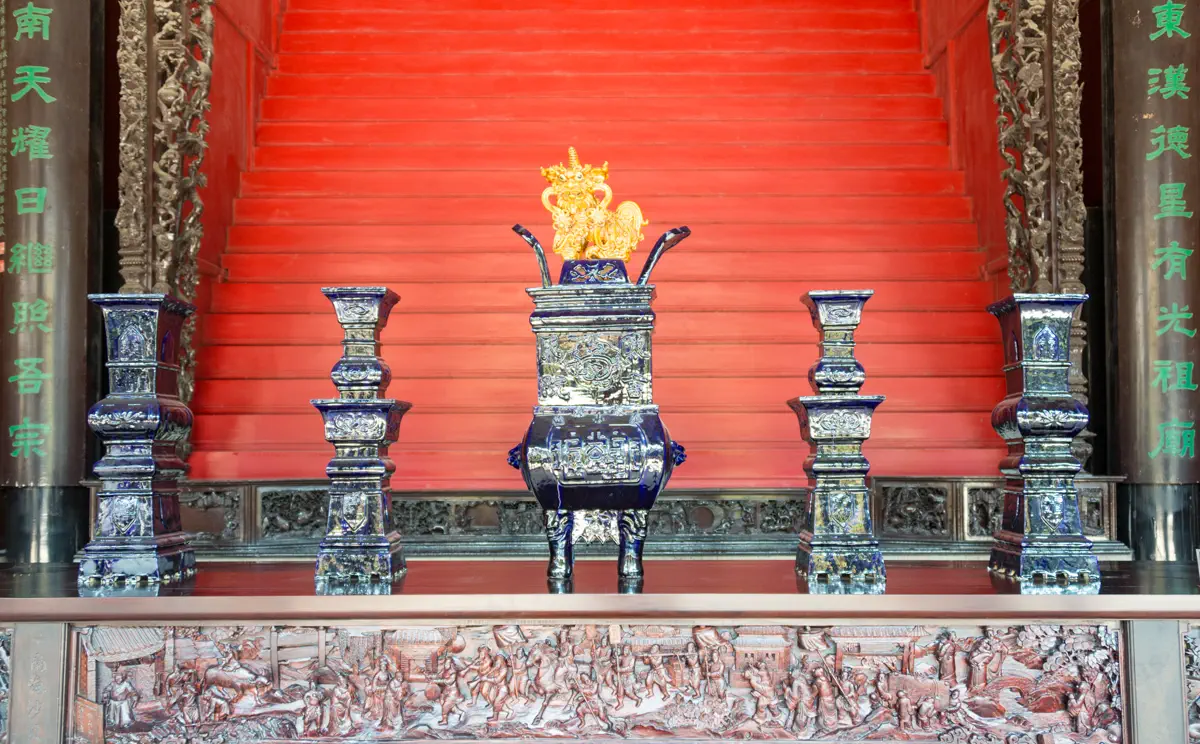Ornate ancestral altar table wood carving in rear hall showing continuous battle scene relief topped with blue glazed bronze incense burner and vases