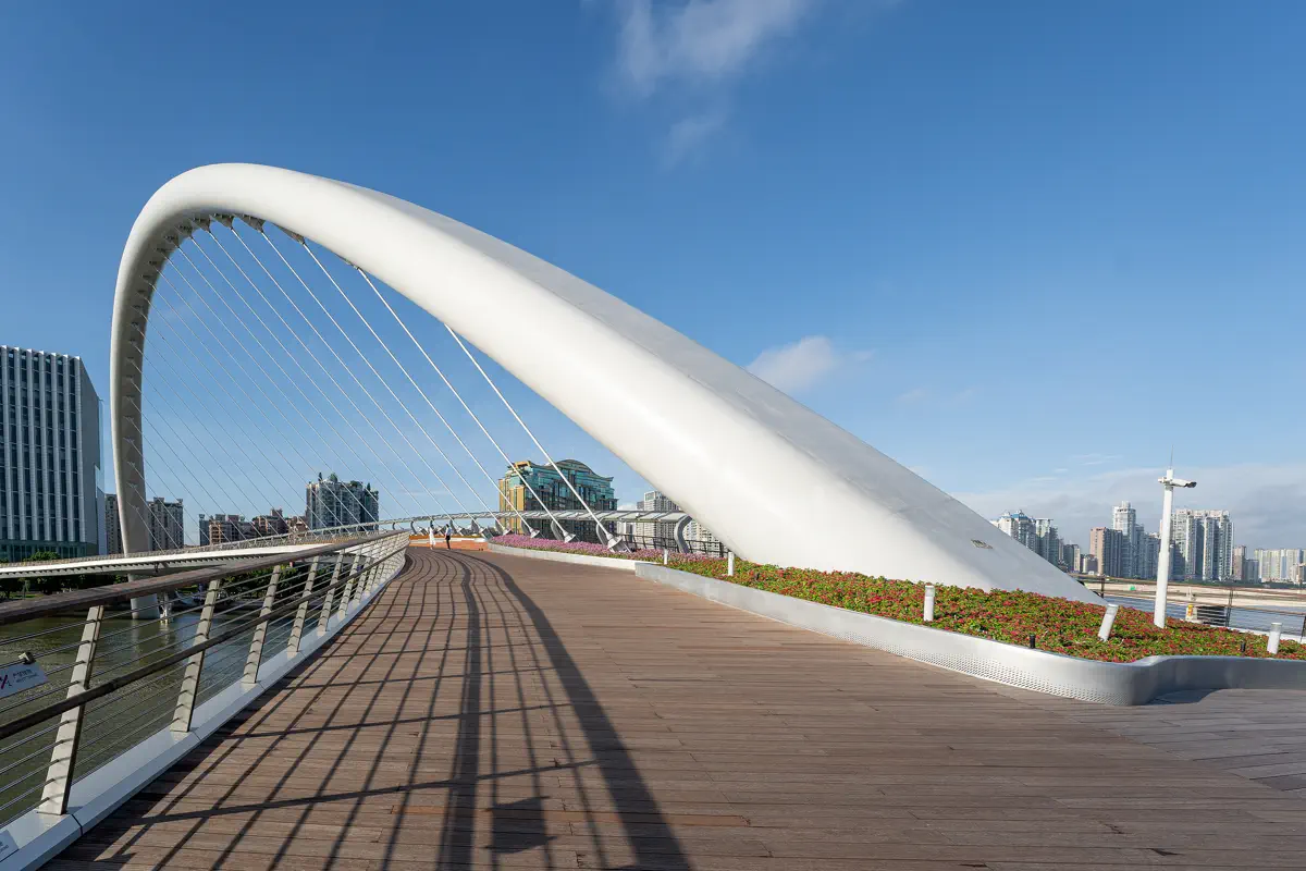Close-up perspective of the wooden pedestrian deck, landscaped flower borders, and dramatic curved white arch of Haixin Bridge