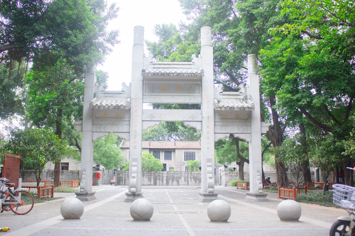 Four-pillar white granite ceremonial archway of Julong Village framed by mature banyan trees