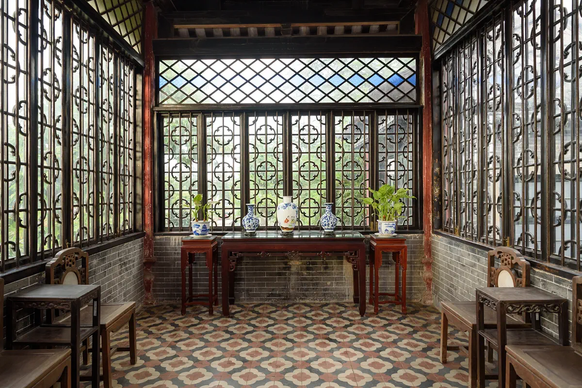 Interior of Linglong Water Pavilion showing intricate wood lattice windows, colorful Manchurian glass, rosewood side tables, and vintage European geometric floor tiles