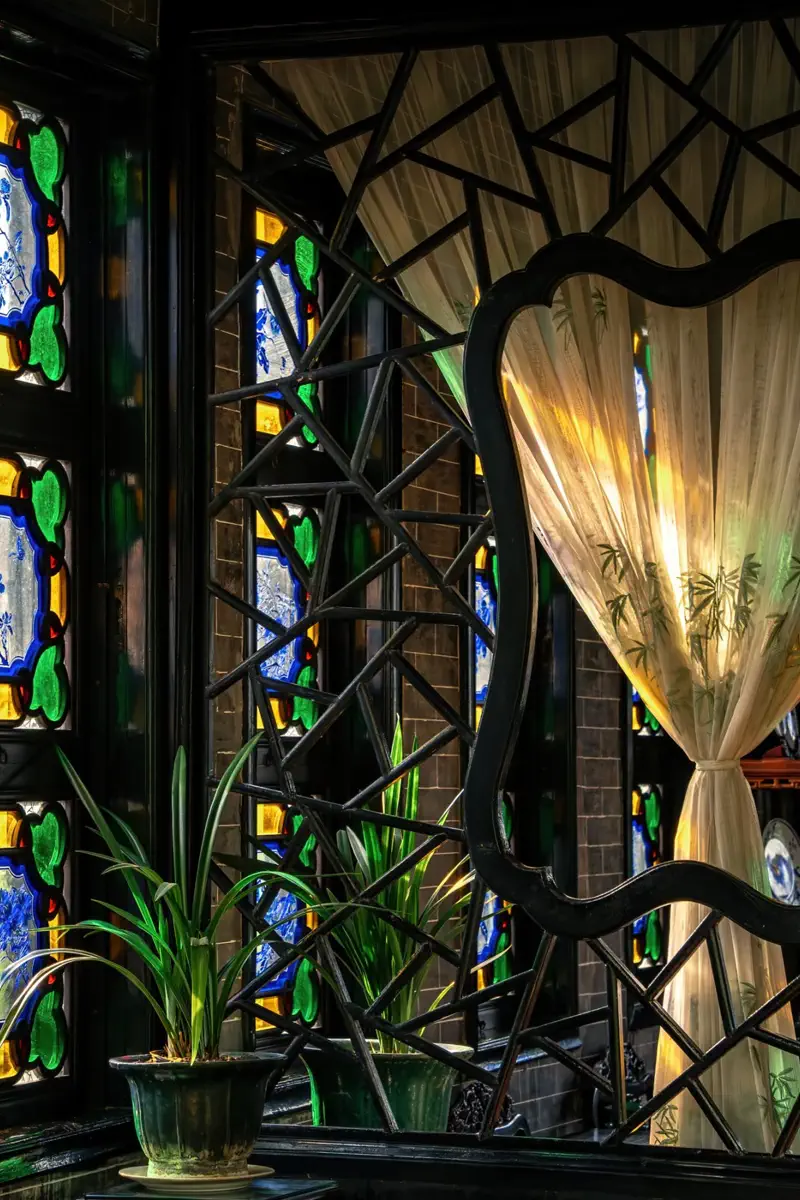 Atmospheric interior in Yuyuan showing geometric wood lattice screens, sunlight streaming through colored glass, and sheer white curtains beside potted orchids